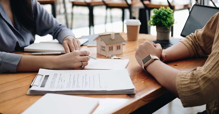 A couple reviews paperwork with a clipboard while standing in the living room of a house with moving boxes nearby.